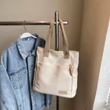 Cartera tipo shopper color beige con bolsillos laterales y un bolsillo frontal con tirador de cordón trenzado. Cuenta con asas superiores de lona.