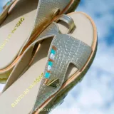 Sandalia plana de cuero metalizado dorado con textura de cocodrilo, diseño cruzado con sujetador para el dedo y suela baja.