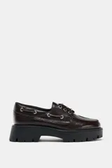 Mocasín de cuero marrón con diseño tipo náutico, presenta ojales metálicos laterales con cordones y suela de plataforma dentada en color negro.
