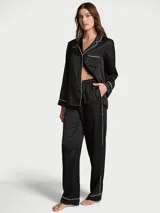 Pijama largo de satín negro, compuesto por una camisa de manga larga con botones, cuello con muesca, bolsillo en el pecho y ribete blanco, y un pantalón largo a juego con cintura elástica.