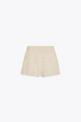 Shorts color beige confeccionados en hilatura con lino, de tiro alto. Presentan bolsillos delanteros, falsos bolsillos traseros con solapa y pinzas delanteras. Cierre frontal con cremallera y gancho metálico.