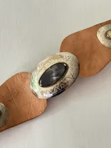 Cinto tipo faja de cuero color marrón, con diseño de piezas de cuero unidas y apliques metálicos plateados grabados con detalles geométricos y una piedra central oscura.