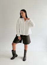 Blusa blanca de corte holgado con cuello mao, botones frontales y detalles bordados en el pecho. Presenta mangas largas con puños con volados.