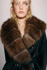 Trench corto Zara de efecto piel color negro, con cuello de solapa y aplicación de pelo sintético a contraste en color marrón. Cuenta con manga larga con trabillas en los puños, bolsillos delanteros y cinturón ajustable del mismo tejido.