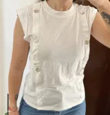 Musculosa Sfera de color blanco, sin mangas y con cuello redondo, destacada por tiras verticales frontales adornadas con botones nacarados.
