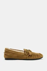 Mocasín de mujer confeccionado en serraje color tostado, con detalle de fruncido y borlas en la parte delantera. Presenta un interior forrado en tejido efecto borreguito y suela plana.
