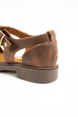 Sandalias tipo cangrejera de cuero color marrón, con diseño de tiras cruzadas, talón cerrado y tira ajustable con hebilla metálica y detalle de estrella grabada en el lateral. Suela plana con pequeño tacón.