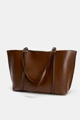 Bolso tipo shopper de color marrón, con diseño minimalista, doble asa de hombro y cierre magnético. Incluye un colgante decorativo con detalle de cinta en tonos claros y un bolsillo interior amplio.