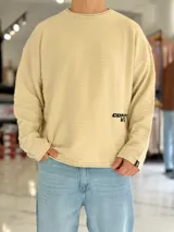Sweater de punto liviano color beige, con textura marcada, cuello redondo y mangas largas de corte relajado, con pequeño estampado tipográfico negro en el lateral inferior.