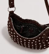 Bolso de hombro estilo hobo en color marrón oscuro, confeccionado en gamuza con diseño de tachas metálicas plateadas distribuidas por toda la superficie. Cuenta con un asa superior fija y cierre superior con cremallera.
