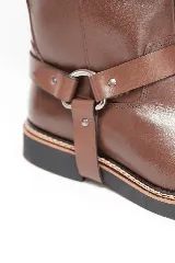Bota de caña alta de cuero marrón, con diseño urbano y líneas limpias. Cuenta con una correa decorativa en el tobillo con argolla metálica y remaches plateados, suela de goma con costura reforzada y tirador trasero superior.
