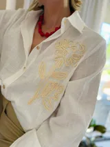 Camisa de manga larga en color blanco, confeccionada en tejido con textura. Presenta un diseño con bordado artesanal en relieve de color crema sobre uno de los hombros y parte del pecho, con cierre frontal de botones.
