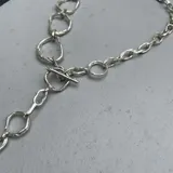 Collar de cadena con eslabones irregulares de diseño artesanal, con cierre frontal tipo barra y aro. La pieza permite diversas formas de uso, incluyendo como collar, pulsera o accesorio de espalda.