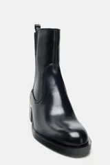 Botín de cuero sintético negro con caña elástica lateral, tirador trasero y detalle de aplique metálico dorado en la base del tacón cuadrado.