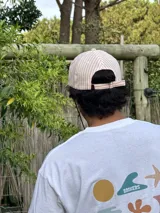 Gorro de visera curva con diseño a rayas verticales en tonos rosado pálido y blanco. La corona tiene un bordado en azul claro con texto. La visera es de color beige claro. Incluye un cordón negro ajustable para colgar del cuello.