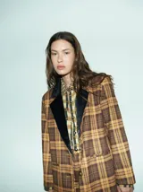 Blazer de corte oversize con estampado de cuadros en tonos marrones y ocres. Presenta solapas de terciopelo negro, cierre frontal con dos botones y bolsillos delanteros. El interior está forrado en satén.