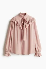 Blusa rosada de manga larga con cuello alto y lazo para atar. Presenta ribetes de volados que cruzan el pecho y puños con elástico fruncido.