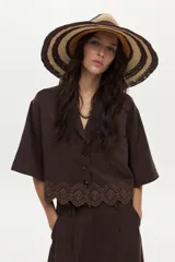 Camisa de manga corta color marrón, con cuello solapa, cierre frontal de botones, bolsillo en el pecho y detalle de bordado decorativo en el ruedo con terminación ondulada.