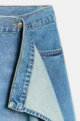 Falda de tiro medio confeccionada en denim, con trabillas, cinco bolsillos, cierre cruzado con botones y bajo acabado asimétrico.