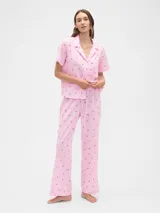 Pijama de dos piezas confeccionado en popelina, compuesto por una camisa de manga corta con cuello solapa y cierre frontal de botones, y un pantalón largo de corte recto con cintura elástica. Presenta un estampado integral de cerezas sobre fondo rosado.