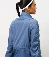 Campera deportiva tipo parka de microfibra en color azul, con cuello alto, cierre frontal por cremallera y mangas largas con puños elásticos. Presenta un diseño con fruncido lateral ajustable y corte amplio.