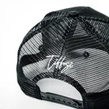 Gorra tipo trucker color negro, con panel frontal rígido y parte trasera de malla. Presenta un logo bordado en relieve de color blanco en el frente.