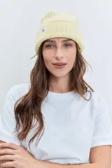 Gorro tipo beanie de punto grueso color amarillo pastel, con vuelta en el borde y etiqueta pequeña con logo en el frente.