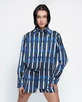 Camisa de corte holgado confeccionada en crepé fino, con estampado geométrico de rayas verticales en tonos azul, blanco y negro. Presenta cuello clásico de tamaño ligeramente ampliado, cierre frontal con botones, mangas largas con pinzas laterales y puños anchos.