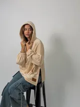 Canguro de plush color beige con capucha, bolsillo frontal tipo canguro, mangas globo con puños fruncidos y tajos laterales con tiras ajustables.