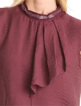 Blusa sin mangas color blanco, confeccionada en crepe con acabado satinado. Presenta un cuello alto decorado con mostacillas y una sobrecapa drapeada en el frente. La espalda cuenta con una abertura tipo gota y cierre de botón.