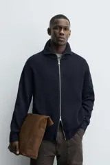 Chaqueta de punto con textura acanalada, cuello subido y cierre frontal completo con cremallera metálica. Presenta un corte relajado y acabados en rib en puños y ruedo.