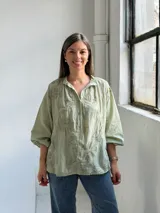 Camisa verde claro de corte oversize, con mangas tres cuartos abullonadas, escote en V con cuello camisero y cierre frontal parcial de botones. Cuenta con delicados bordados florales tono sobre tono distribuidos por toda la prenda.
