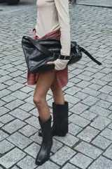 Bolso tipo maxi saca de cuero negro, con asa de hombro ajustable adornada con ojales metálicos plateados. Presenta cierre principal de cremallera y bolsillo interior con cierre.