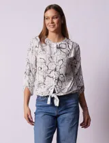 Blusa blanca estampada con flores negras, cuello redondo, abertura con botones y mangas 3/4 con frunce. Tiene terminación con nudo en el ruedo.