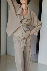 Camisa de lino color tostado con cuello subido, manga larga con puños elásticos y volantes, cintura ajustable con lazadas laterales y cierre frontal cruzado con doble botonadura.