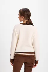 Cardigan de punto color crudo con detalles en marrón oscuro. Presenta cuello redondo, cierre frontal con botones tipo alamares y puños y ruedo acanalados con rayas horizontales. Incluye un pequeño aplique bordado en la manga.