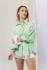 Camisa verde pastel de algodón, corte oversize, con cuello camisero, cierre con botones, mangas largas con puños con volados de encaje blanco, dos bolsillos frontales con solapa y ruedo irregular. 