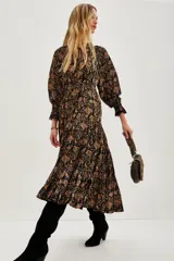 Vestido midi de corte fluido con estampado paisley en tonos oscuros, ocres y rosados. Presenta cuello redondo con lazo de ajuste, mangas largas abullonadas con puños elásticos y falda con volados en el ruedo.
