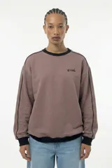 Buzo de cuello redondo en color marrón topo, con diseño de corte relajado. Presenta detalles de piping negro que recorren los hombros y las mangas, además de puños, cuello y cintura en tejido rib negro. Incluye el logo de la marca estampado en el pecho.