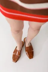 Zueco de cuero color marrón, estilo mule, con puntera cuadrada y tira ancha adornada con flecos. Suela plana.