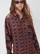 Camisa de seda con cuello camisero, manga larga y bolsillo frontal. Presenta un diseño estampado geométrico de cuadrados en tonos marrones y azul oscuro.