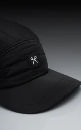 Gorra negra de estilo 5 paneles, con tejido acolchado o con costuras horizontales y visera plana. Presenta un pequeño parche cuadrado negro con un logo blanco bordado en forma de 'X' o lazo.