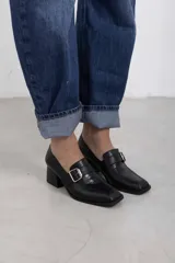 Mocasines negros de cuero con taco bloque, punta cuadrada y tira frontal con hebilla metálica y detalle texturizado.