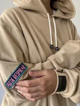 Hoodie oversize color beige, con capucha, cordones ajustables y puños elásticos. Presenta un diseño liso con una pequeña etiqueta de marca en la manga.