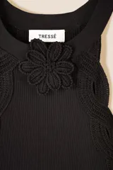 Vestido largo de punto acanalado en color negro, con diseño de sisa americana y cuello redondo decorado con una flor de crochet hecha a mano. Presenta un ribete de trenza de crochet y una abertura lateral en la parte inferior.