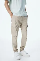 Pantalón cargo color beige de corte slim fit, confeccionado en tejido de algodón. Cuenta con cintura elástica ajustable con cordón, bolsillos al bies, bolsillos laterales y traseros con solapa y botones a presión ocultos, costuras y pinzas en las rodillas.