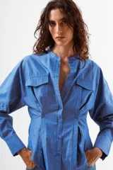 Camisa azul de algodón con finas rayas blancas verticales, cuello mao, manga larga y detalle de pinzas y botones en la zona de la cintura. Posee dos bolsillos de parche con tapa en el pecho.