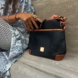 Cartera tipo morral en cuero sintético negro con detalles en cuero marrón y cierres dorados. Cuenta con bolsillos frontales con cierre y correa ajustable.