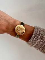 Pulsera con banda de terciopelo verde oliva y dije circular dorado con figura de paloma y rayos.