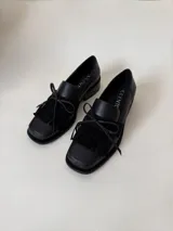 Mocasín de cuero marrón con textura granulada, diseño de horma cuadrada, detalle de flecos y lazo decorativo en el empeine, montado sobre una suela y taco bajo de color negro.